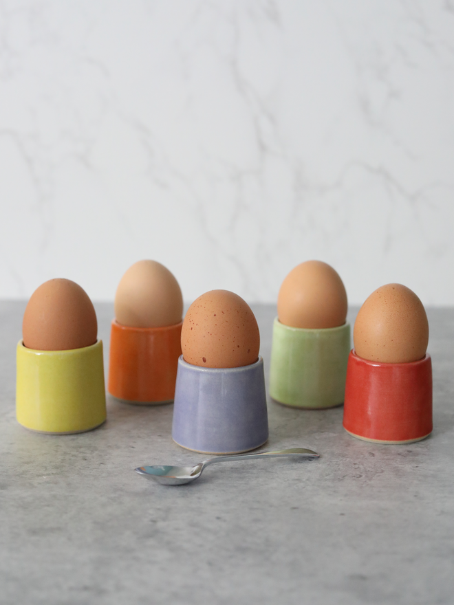 Five handmade ceramic egg cups each holding a brown egg, arranged on a grey marble surface in yellow, orange, purple, spring green, and cherry red glazes, with a small silver spoon in the foreground and a white marble background