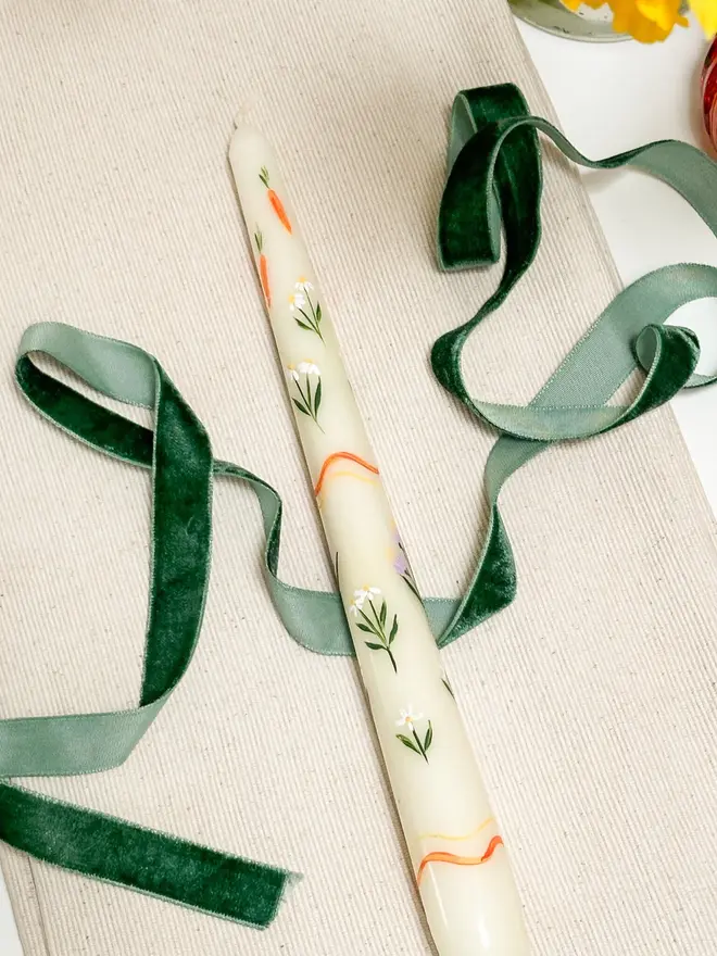 Carrots And Flowers Spring Candle, a spring candle lying on a plain surface accompanied by a green ribbon. It's floral design can be seen wrapping around it. 