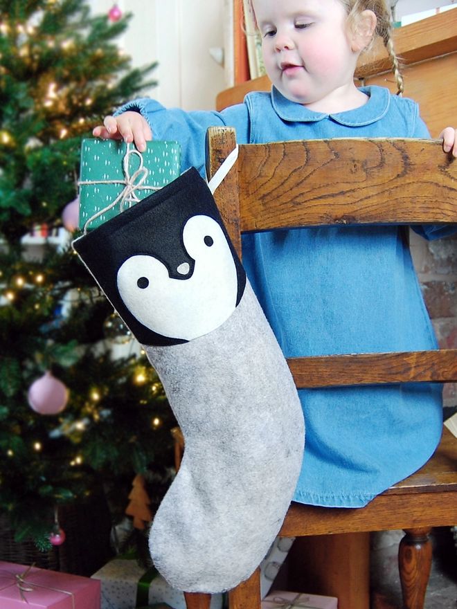 A young girl wearing a denim dress stands in front of a Christmas tree pulls a present out of a handmade felt penguin stocking.