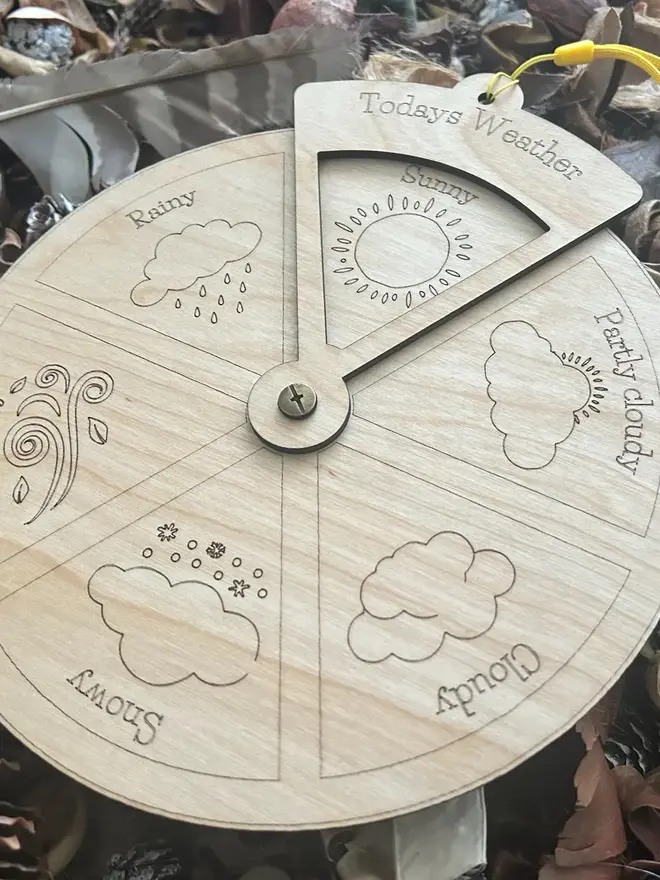 Extra large wooden weather wheel with engraved weather symbols for sunny, cloudy, rainy, windy, snowy, and partly cloudy, shown resting among pinecones and natural materials.