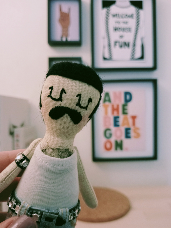 Close up of Freddie mercury mini decorative icon doll held up in front of a mini gallery wall of music themed prints Freddie is wearing his iconic live aid era outfit of white vest tight bleach jeans and studded belt and arm band