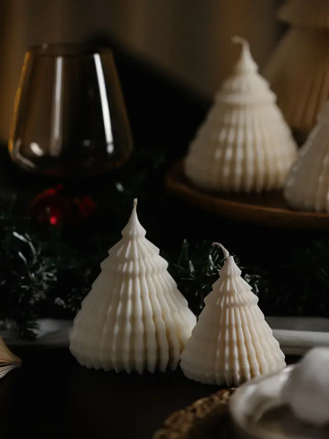 Origami Christmas Tree Candles (Set Of 2). Two unlit cream tree candles with finely ridged tiers, styled on a dark table with pine garland, a wine glass and a blurred tray of matching candles behind. The minimalist handmade design makes a thoughtful Christmas gift or place-setting decoration.