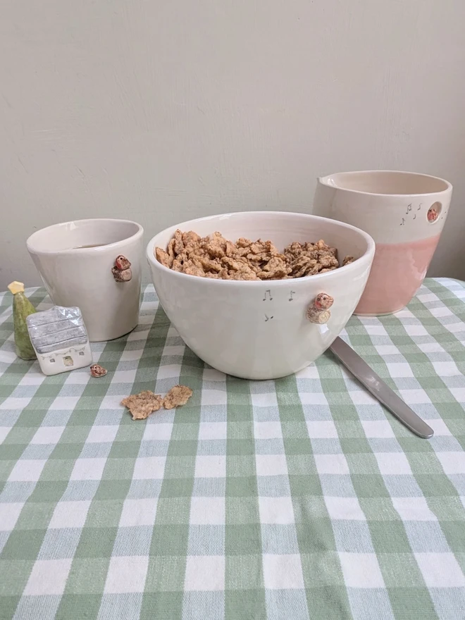 white ceramic bowl with a tiny modelled robin perching on a modelled branch with musical notes, the bowl in full of breakfast cereal and there is a pink jug with a robin and espresso robin cup next to the bowl sitting on a green gingham tablecloth