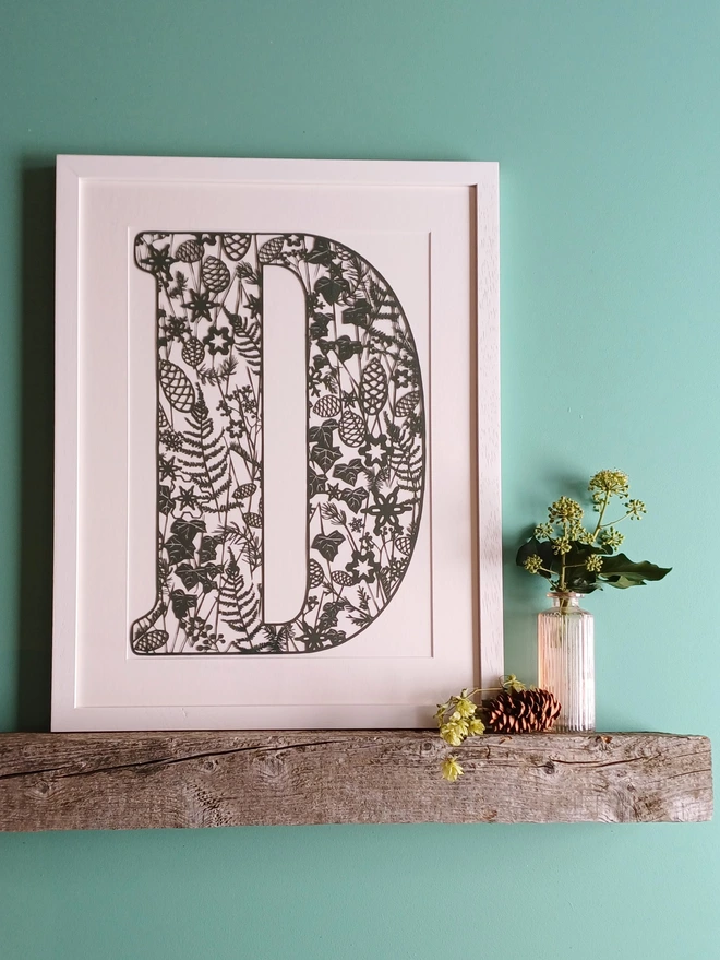 Papercut Letter D in dark green with winter botannical details. Displayed in narrow white wooden frame with off white mount. 