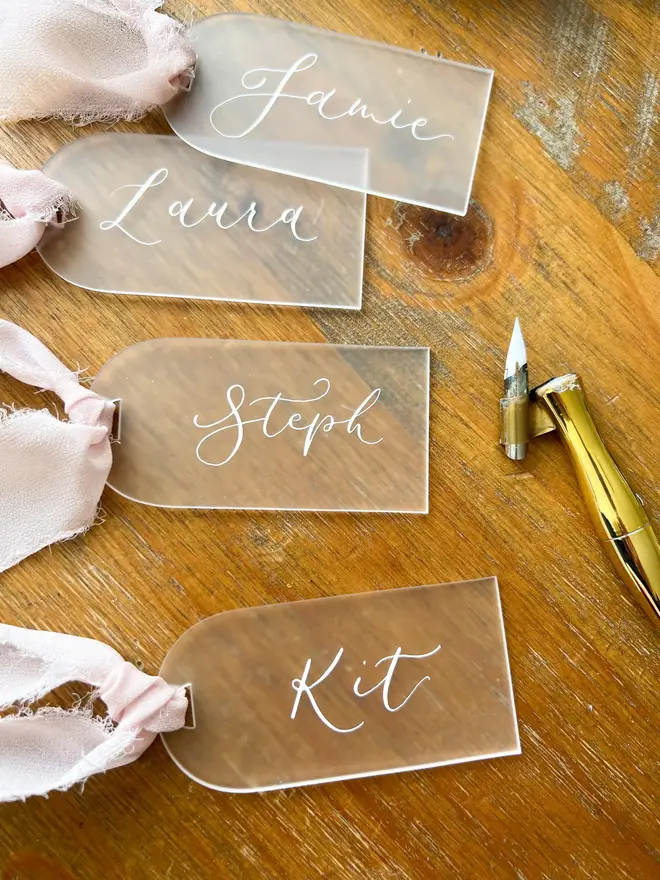 Personalised Calligraphy Frosted Acrylic Place Cards, frosted acrylic place cards with pink ribbons grouped together on a wooden table. 