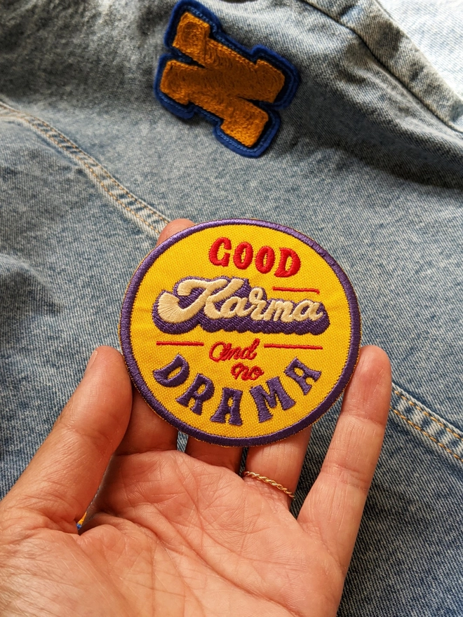 Yellow, Blue & Red round typographic patch reading 'Good Karma and No Drama'. The word Karma is written in a thick cursive lettering style in white, with a blue drop shadow, while the additional type is written in a retro style capital lettering & a smaller red cursive making up a fun retro style fabric patch. 