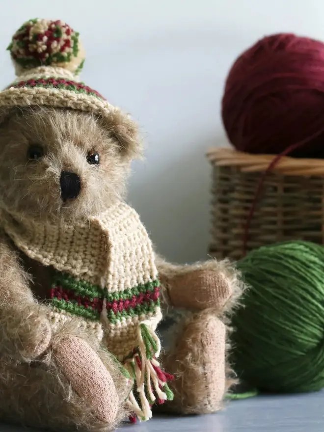 Brown teddy bear sitting upright against a white background among round balls of wool, wearing a knitted beige hat and scarf with green and burgundy details.
