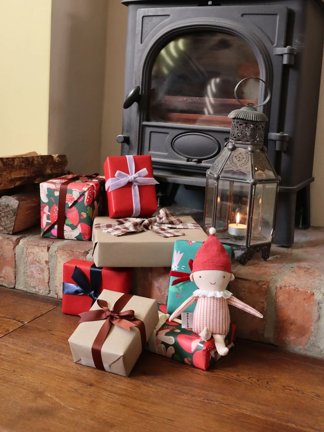 handmade elf doll in stripe top and red  hat with a name embroidered, in front of a pile of gift boxes and fireplace. 
