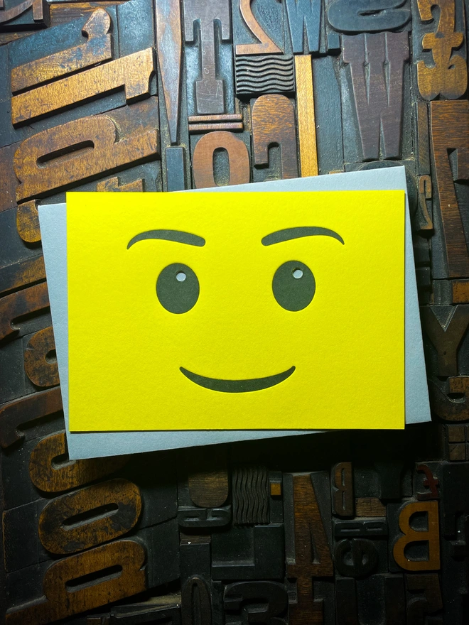 A bright yellow letterpress card with a smiling face emoji is placed on top of a grey envelope. The background consists of various wooden printing letters in different orientations and sizes.