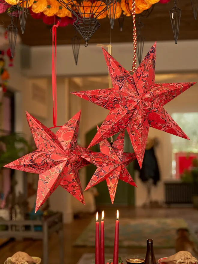 Red marbled paper hanging star decorations in various sizes (small, medium & large), hung above a busy Christmas tablescape from red ribbon
