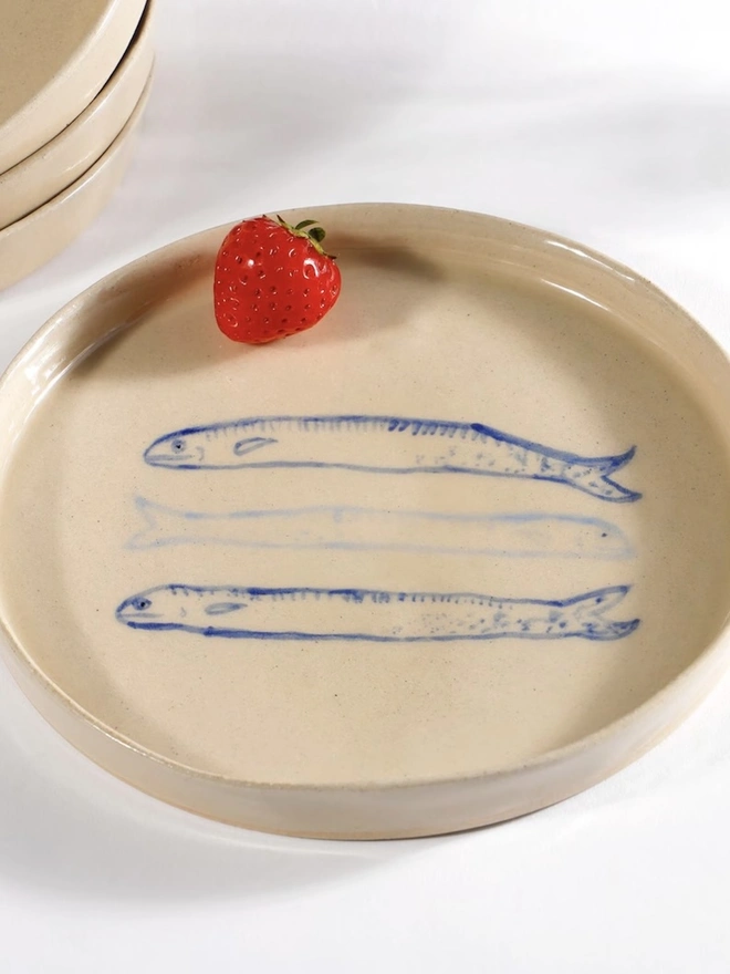 Handmade Ceramic Side Plate With 3 Blue Sardines Design , a side plate with a fish design sitting on a white table surface, accompanied by a raspberry. 