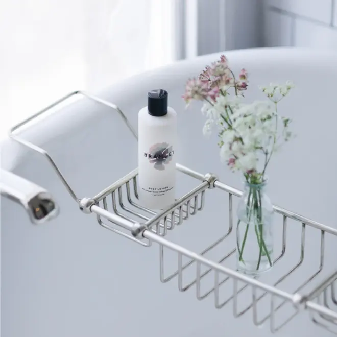 white bottle of lotion with black lid, sitting in a silver bath caddy with a small glass vase of white flowers