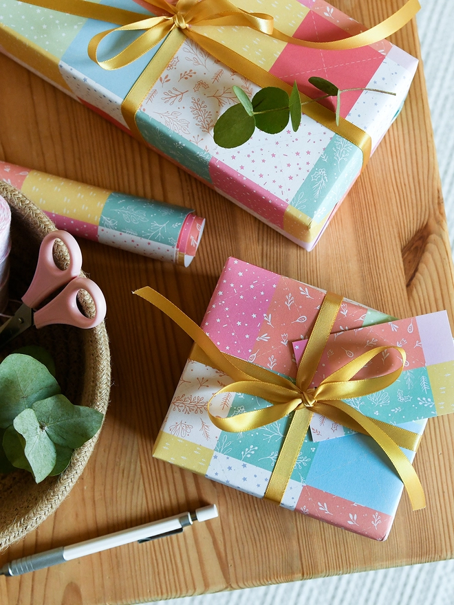 Gifts wrapped in gift wrap with a design of pastel patchwork quilt design is on a wooden table.