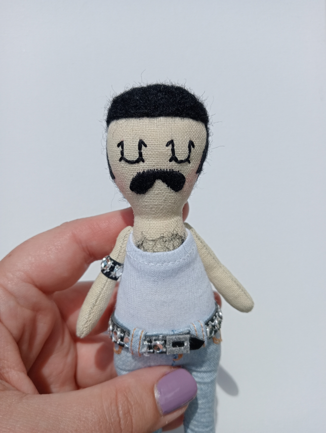 Mini decorative Freddie Mercury doll held in a left hand against a white background Freddie is wearing a white vest tight bleached jeans and black studded belt and arm band
