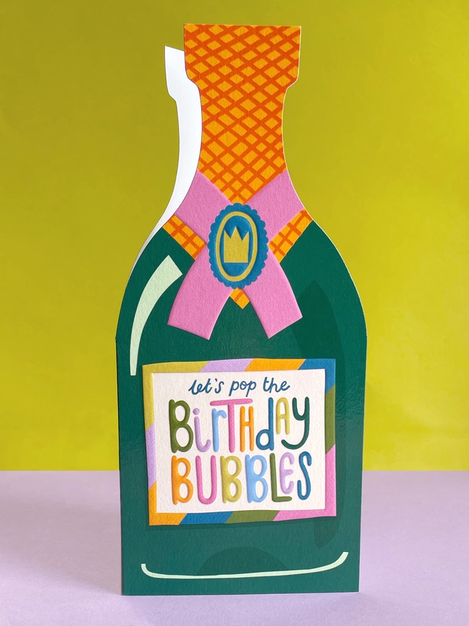 A vibrant die cut birthday card in the shape of a champagne bottle with a rainbow coloured ‘Let’s pop the birthday bubbles’ message