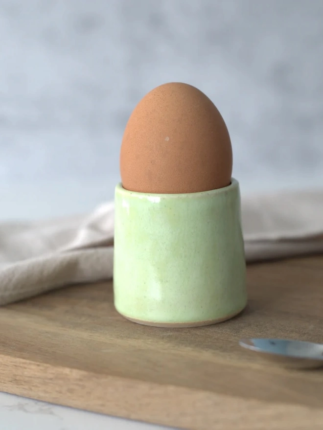 Classic Handmade Egg Cup, a ceramic egg cup sitting on a wooden chopping board against a grey backdrop. It is accompanied by a spoon
