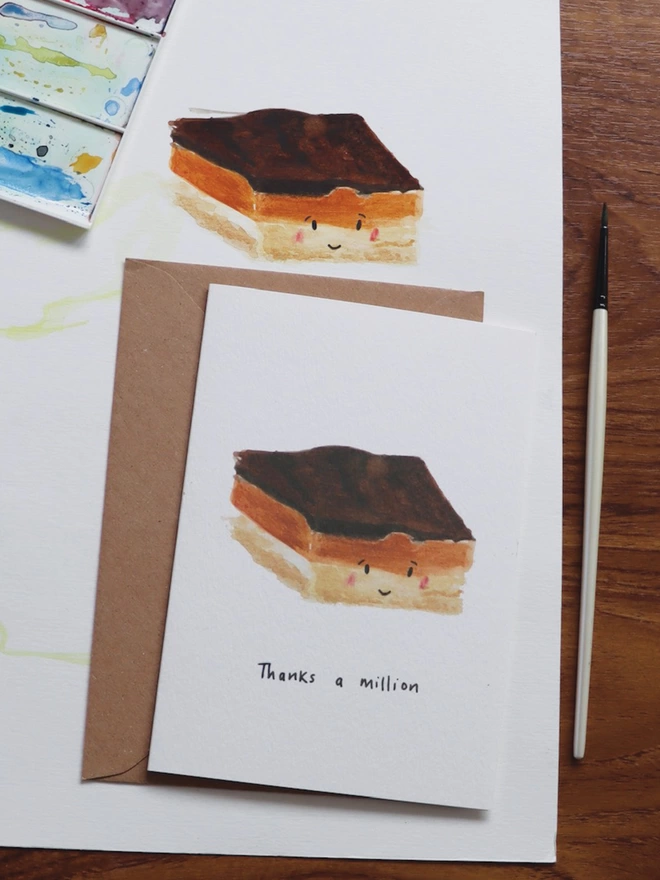 Desk Shot Of The Thanks a Million Millionaire’s Shortbread Thank You Card Sitting Along Side The Original Hand Painted Watercolour Illustration, Paintbrush and Palette