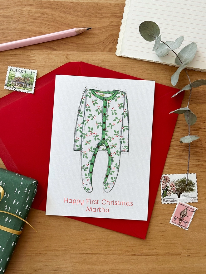 A personalised first Christmas card with an illustrated onesie lays on a red envelope on a wooden desk.