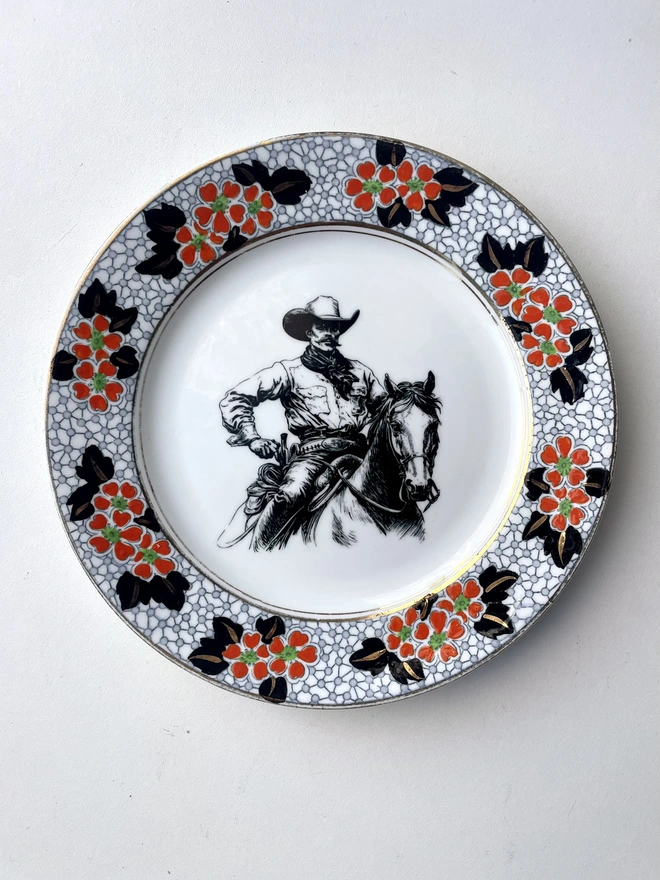 vintage plate hanging on a white wall, it has a decorative border, with a printed vintage illustration of a cowboy in the middle 