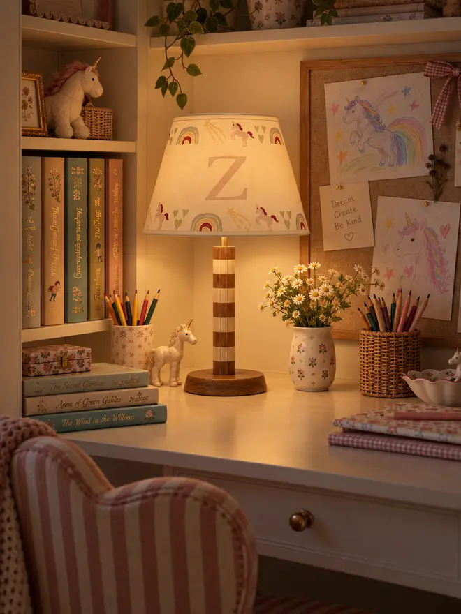 Personalised unicorn lampshade with initial Z glowing warmly on a striped wooden table lamp in a cosy girls’ bedroom study area.