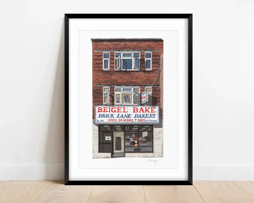 Brick Lane Beigel Bake Giclée Print (Unframed) Shoreditch, a print of a cafe sitting in a black frame on a wooden surface against a plain backdrop. 