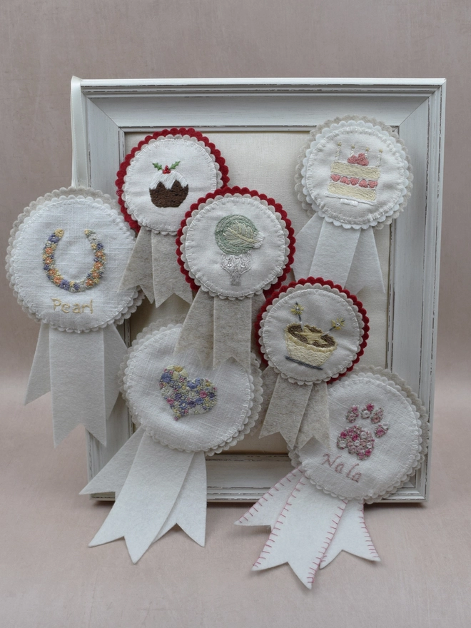 Finished embroideries made-up into felt rosettes, pinned onto a padded frame. Displayed are the kits for Birthday Cake, Horseshoe, Heart, Cat Paw, Christmas Pudding, Brussels Sprout & Mince Pie.