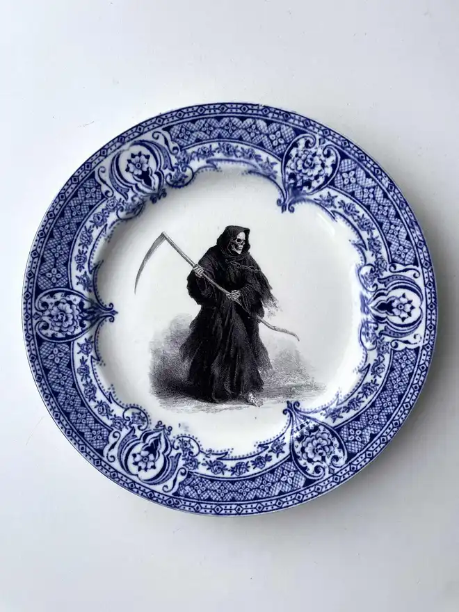 a vintage plate with an ornate border has a vintage illustration in black and white of a grim reaper skeleton in the centre of the plate