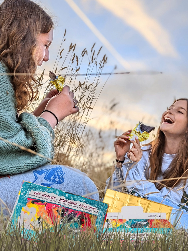 Two happy girls in a field enjoying charity dark chocolate bar wrapped in gold foil 