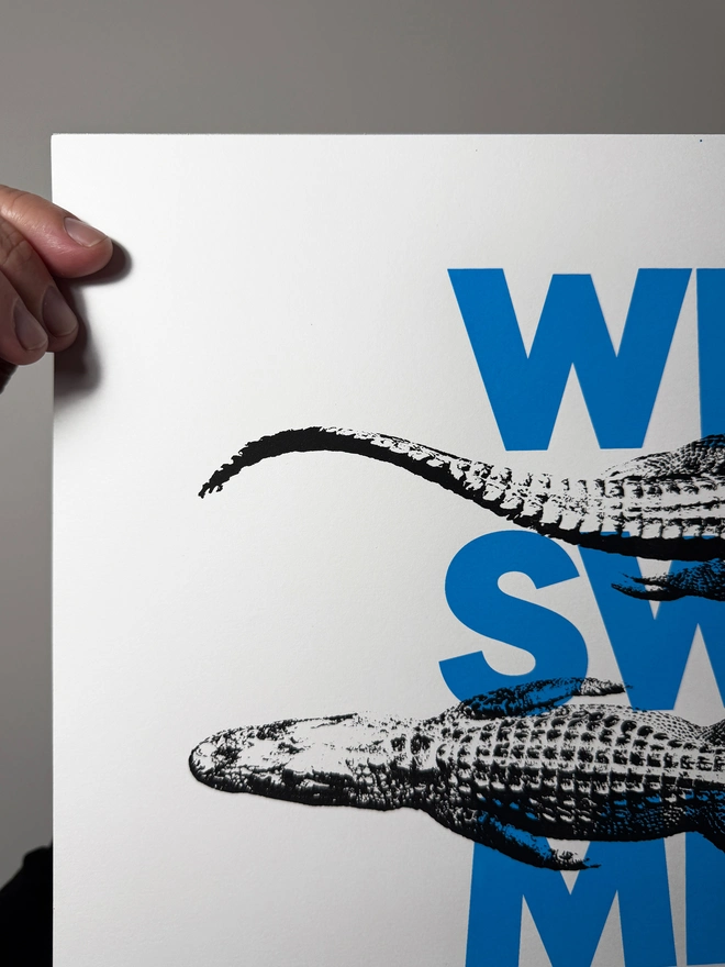 A humorous limited edition A3 screen printed poster. Featuring black alligators over blue 'wild swimming' typography. Showing close up of detail top left.