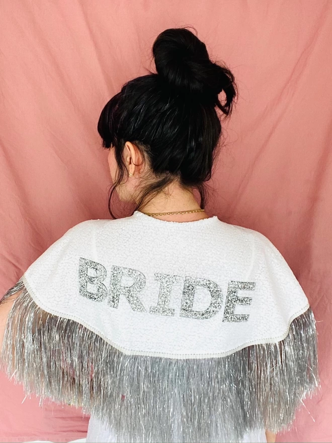 A woman wearing the gretna bride cape. The word 'BRIDE' is written across the cape in silver chunky glitter on top of white sequins. The cape has a silver tinsel fringe along the bottom. 