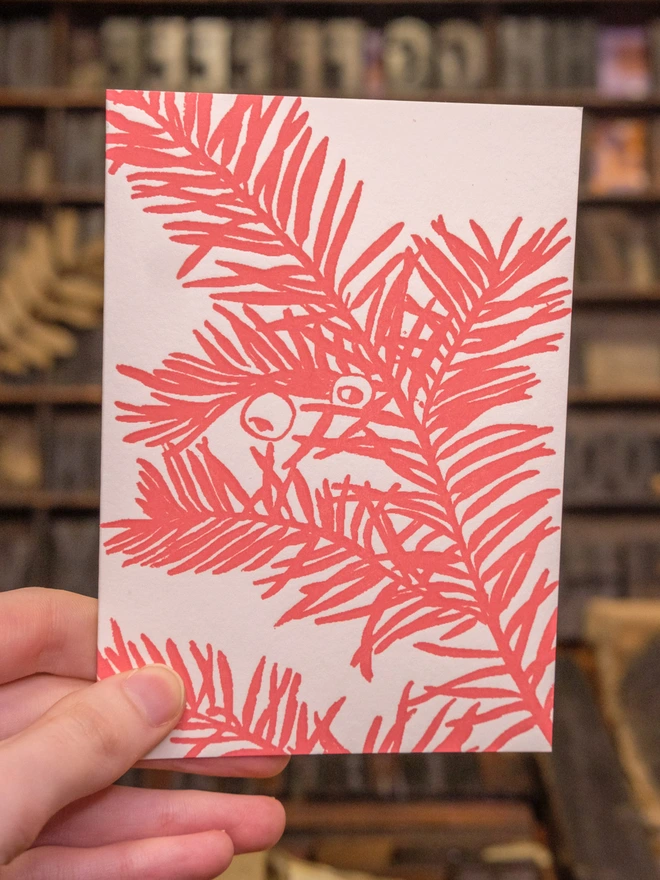 Card held up. Situation shot. Design on front visible. Monochrome red artwork of yew branch 