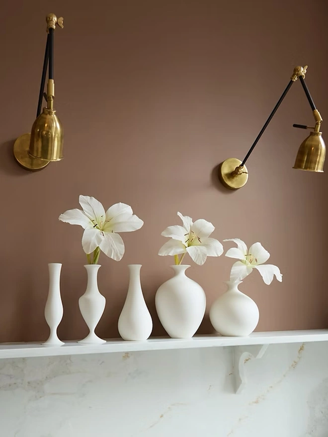 collection of multi shaped white 3d printed vase with white flowers on white shelf against brown wall