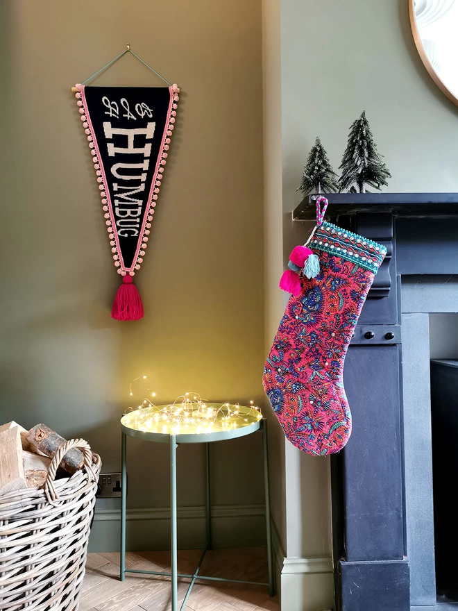 A Bah Humbug banner is hung in a green living room alcove, beside a fireplace. A stocking in complimentary colours hangs from the mantlepiece besides a basket full of firewood and a table glowing with fairy lights.