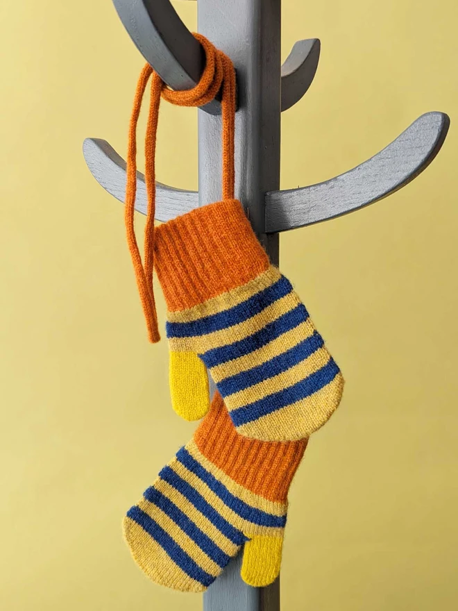 Kids Blue And Green Stripe Mittens On A String Hanging On A Coat Stand In Front Of  A Green Background
