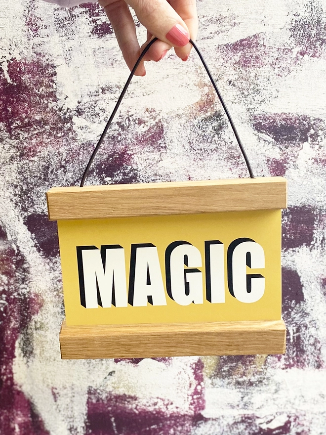 A print in yellow with the word MAGIC framed in a oak hanger, with a waxed cotton cord for hanging being held by a hand with pink nail varnish 