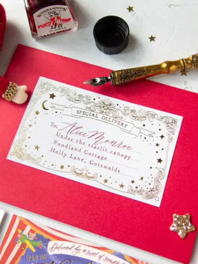 A close-up of a personalised gold foil "Special Delivery" label on a red envelope, addressed "To: Alice Monroe, Under the starlit canopy, Woodland Cottage, Holly Lane, Cotswolds", beside a gold dip pen and a bottle of red ink.