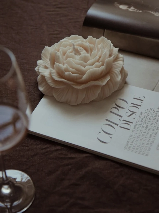 Large White Peony Candle. A cream peony-shaped candle with layers of finely detailed petals, resting on an open magazine beside a wine glass on a brown linen cloth. The realistic floral design makes an elegant decorative piece for a coffee table or bedside.