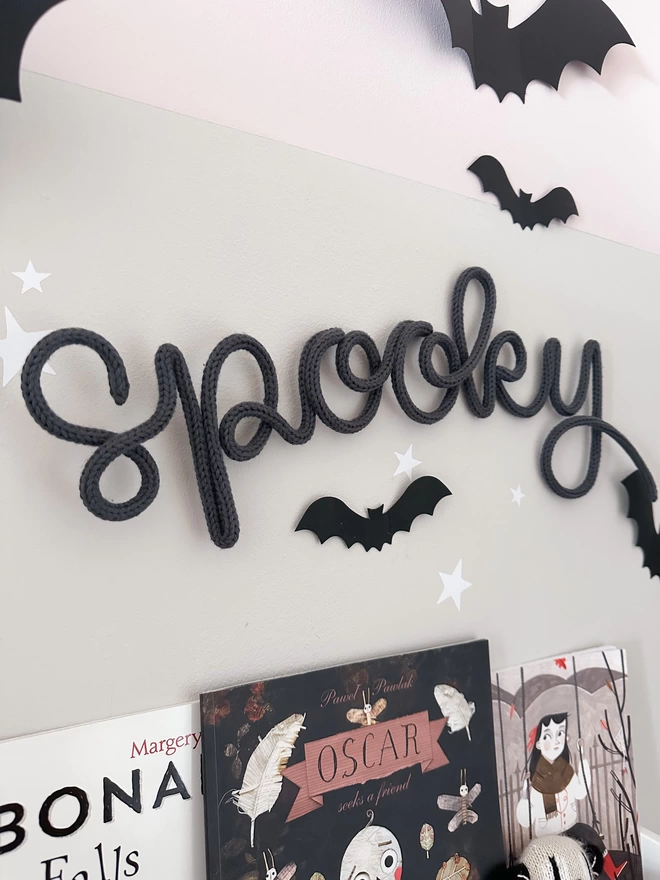 A knitted wire word sign for Halloween - 'spooky' adorned with colour graphite knitted icord hanging above a shelf with kid's halloween books and some paper bat wall decorations. 
