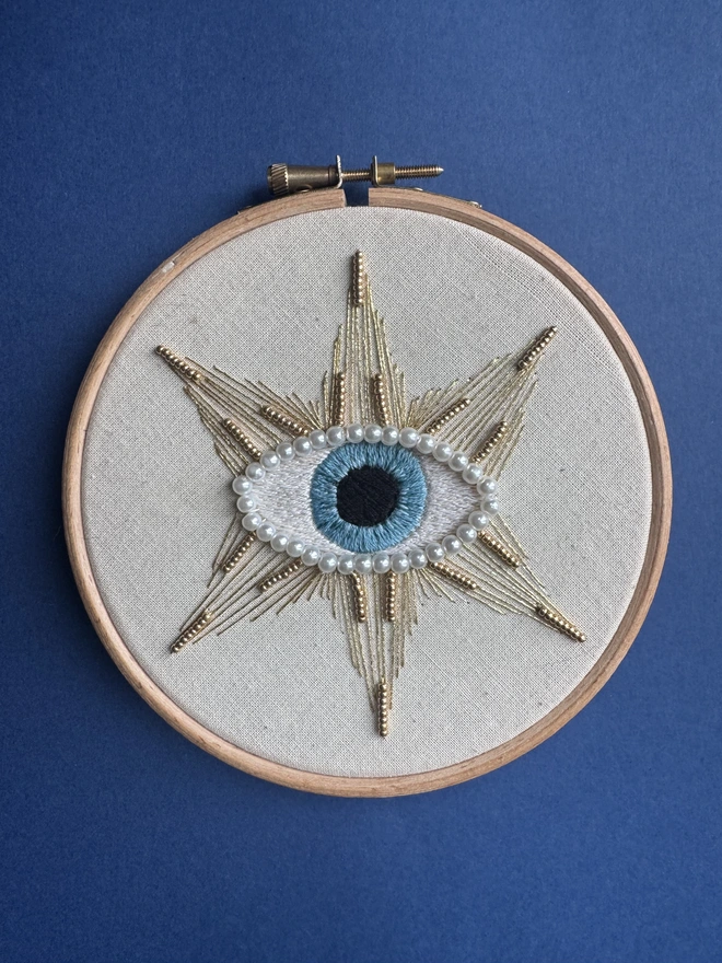An embroidery hoop on a blue background containing a sewn protective eye edged with pearl beads surrounded by a stitched gold star with gold beads