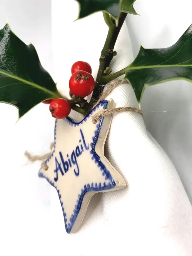 Personalised Ceramic Christmas Star Name Place Markers, a colourful ceramic start sitting on a white surface attached to a holly bush. 