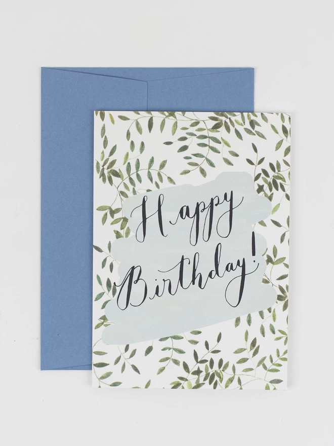 A birthday card with a message written in black calligraphy reading "happy birthday". The card is decorated with delicate vines and comes with a corresponding pale blue envelope.