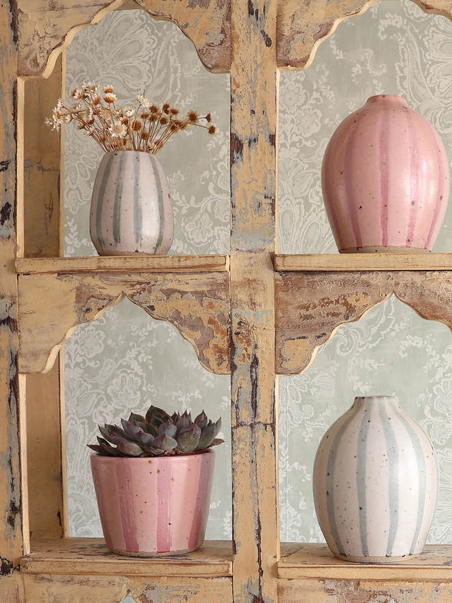 A vintage wooden shelving unit holding a handmade ceramic stoneware vase in blue and white stripe filled with dried flowers, two medium rounded vases - one in a blue and white stripe and one in a pink stripe design – and a handmade ceramic stoneware plant pot with a pink stripe design, holding a succulent plant. 