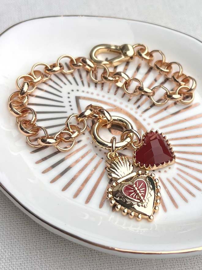 Gold belcher chain bracelet with red heart charm and Frida Kahlo heart charm on a white and gold evil eye dish