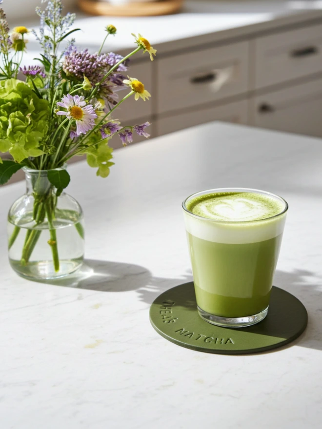 Thea's Matcha stamped on a green coaster, with a matcha drink resting on it. A vase of meadow flowers in the background