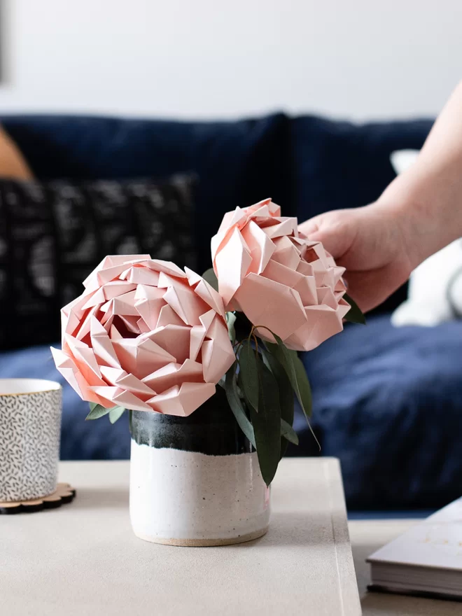 Two blush pink paper peonies on a coffee table in a living room with a hand reaching out to gently touch them.