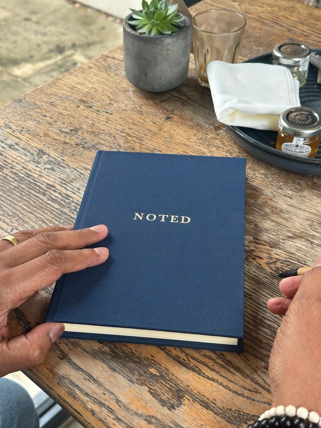 An notebook printed with 'Noted' in gold lettering is held in a man's hands above a wooden table.