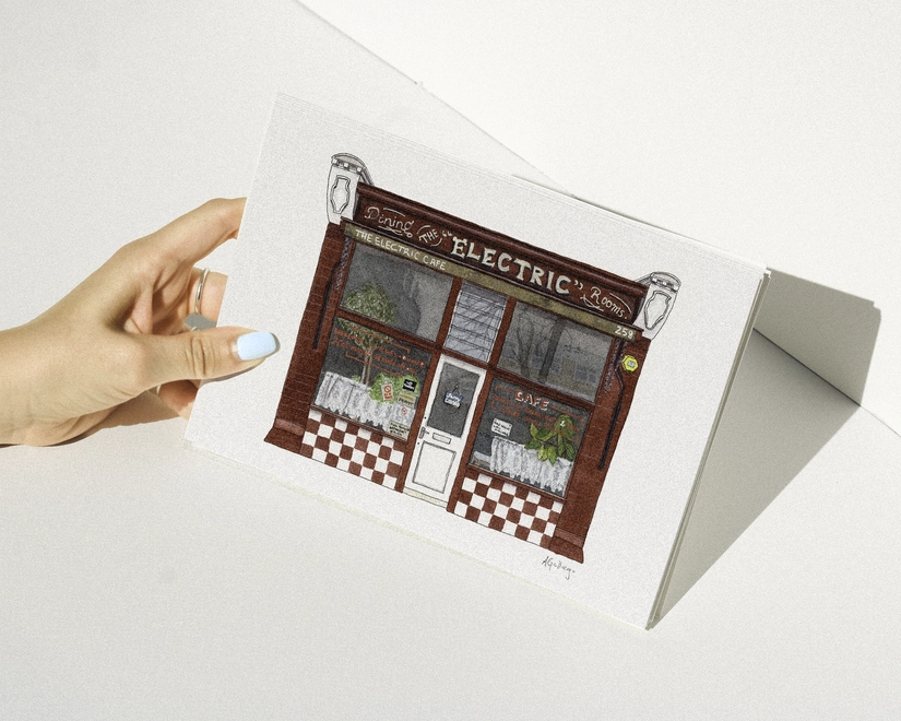 A hand with blue nail polish holding a print featuring an illustration of the red-fronted Electric Cafe with its checkered "Dining Rooms" sign against a white background.