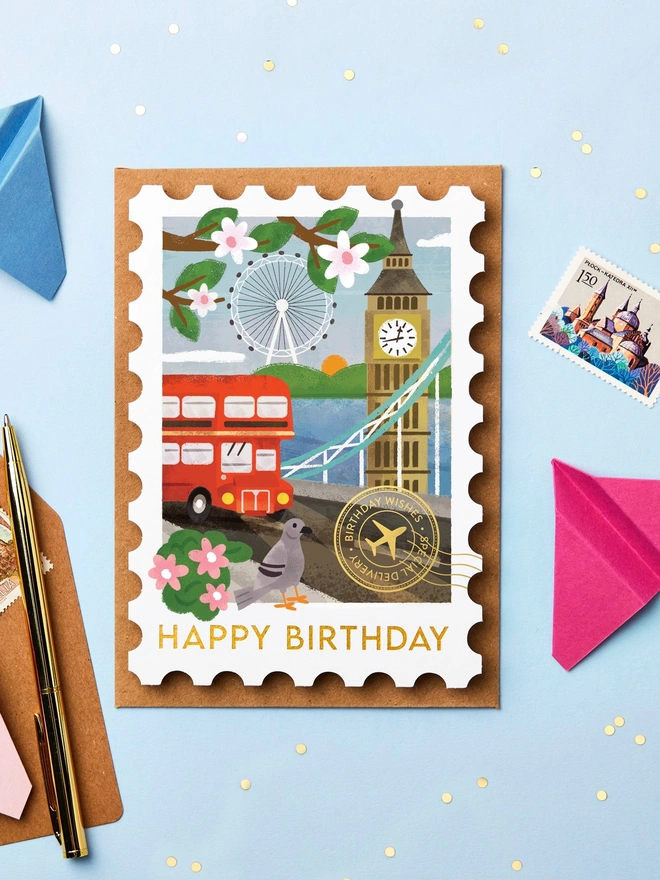 Stamp-shaped greeting card featuring an illustration of London. The card is on a blue background with paper aeroplanes and stationery around it.