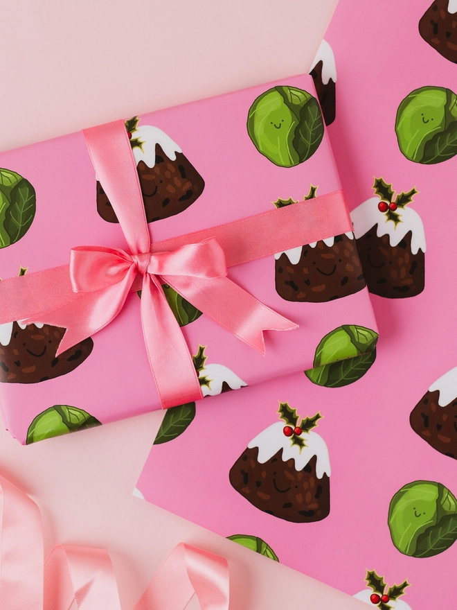 Pink Christmas wrapping paper featuring a repeating pattern of hand-drawn Christmas puddings with white icing and holly and smiling green Brussels sprouts.