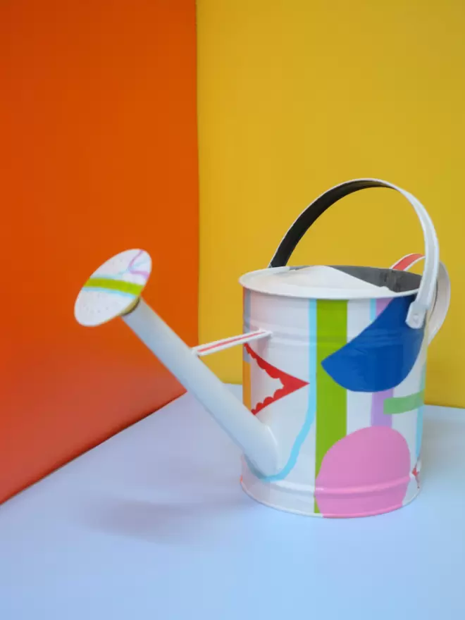 Hand painted watering can by Julie-Anne Pugh. Base colour is white with bring coloured shapes weaving across the body of the can on a yellow and orange background. Photographed from the front.